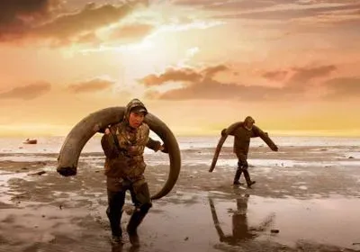 two men carry large tusks over a beach